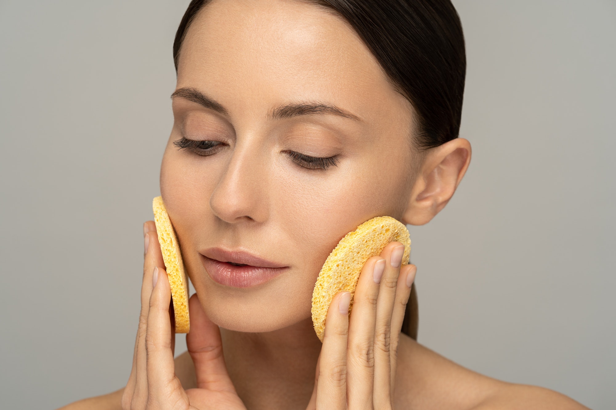 Young woman with nude make-up and naked shoulders cleaning her face with exfoliating sponge isolated